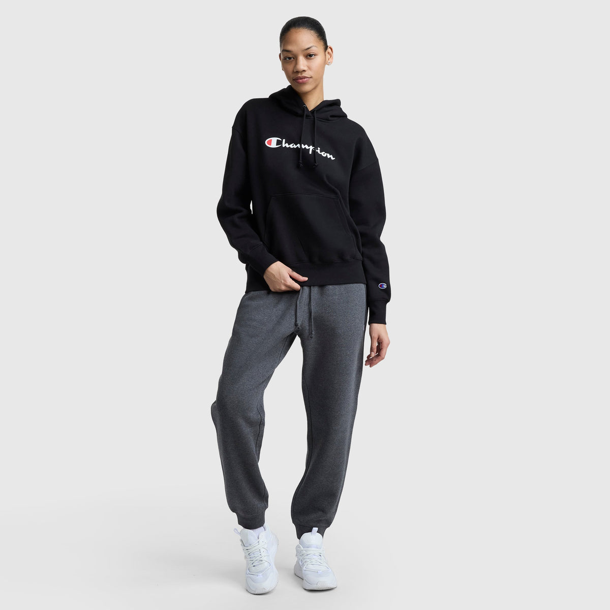 A person poses casually against a plain background, wearing the Champion Powerblend Relaxed Hoodie in black, gray sweatpants, and white sneakers—one hand in their pocket and the other on their hip.