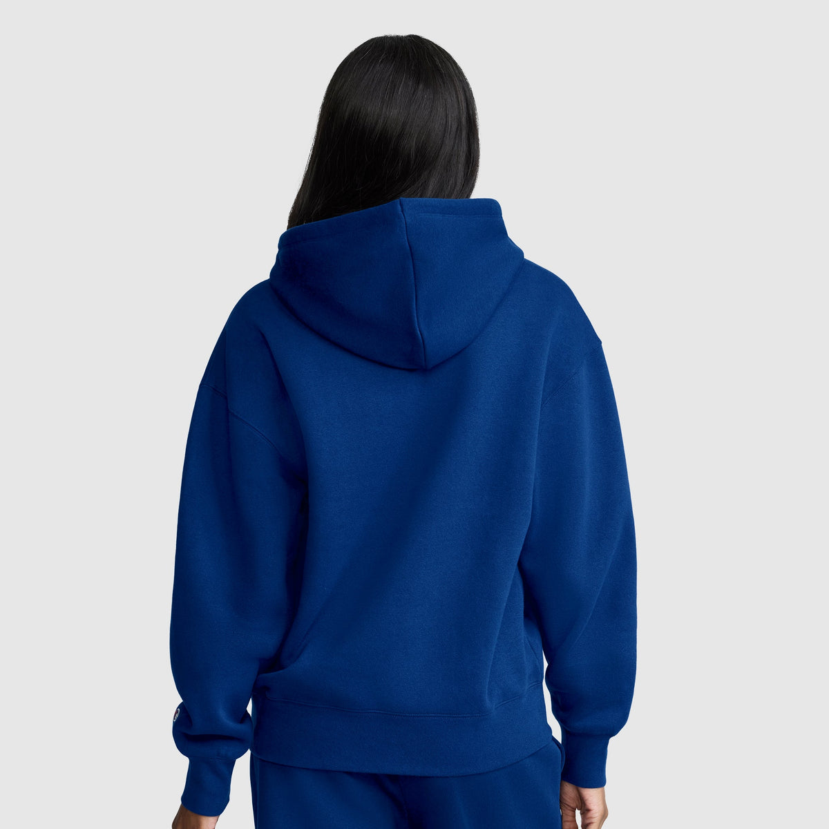 A person with long dark hair, facing away from the camera against a plain light gray background, wears a Champion Women’s Powerblend Relaxed Hoodie, Script Logo and matching blue pants. The midweight fleece of this Champion hoodie provides both comfort and style.
