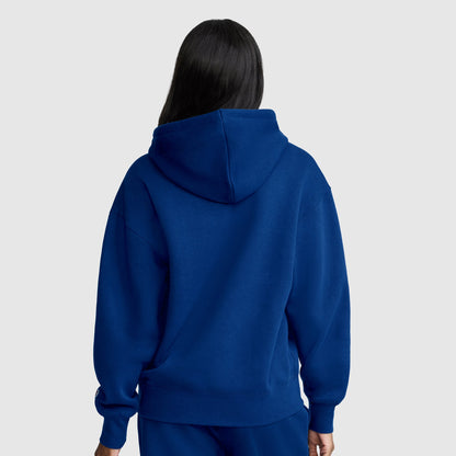 A person with long dark hair, facing away from the camera against a plain light gray background, wears a Champion Women’s Powerblend Relaxed Hoodie, Script Logo and matching blue pants. The midweight fleece of this Champion hoodie provides both comfort and style.
