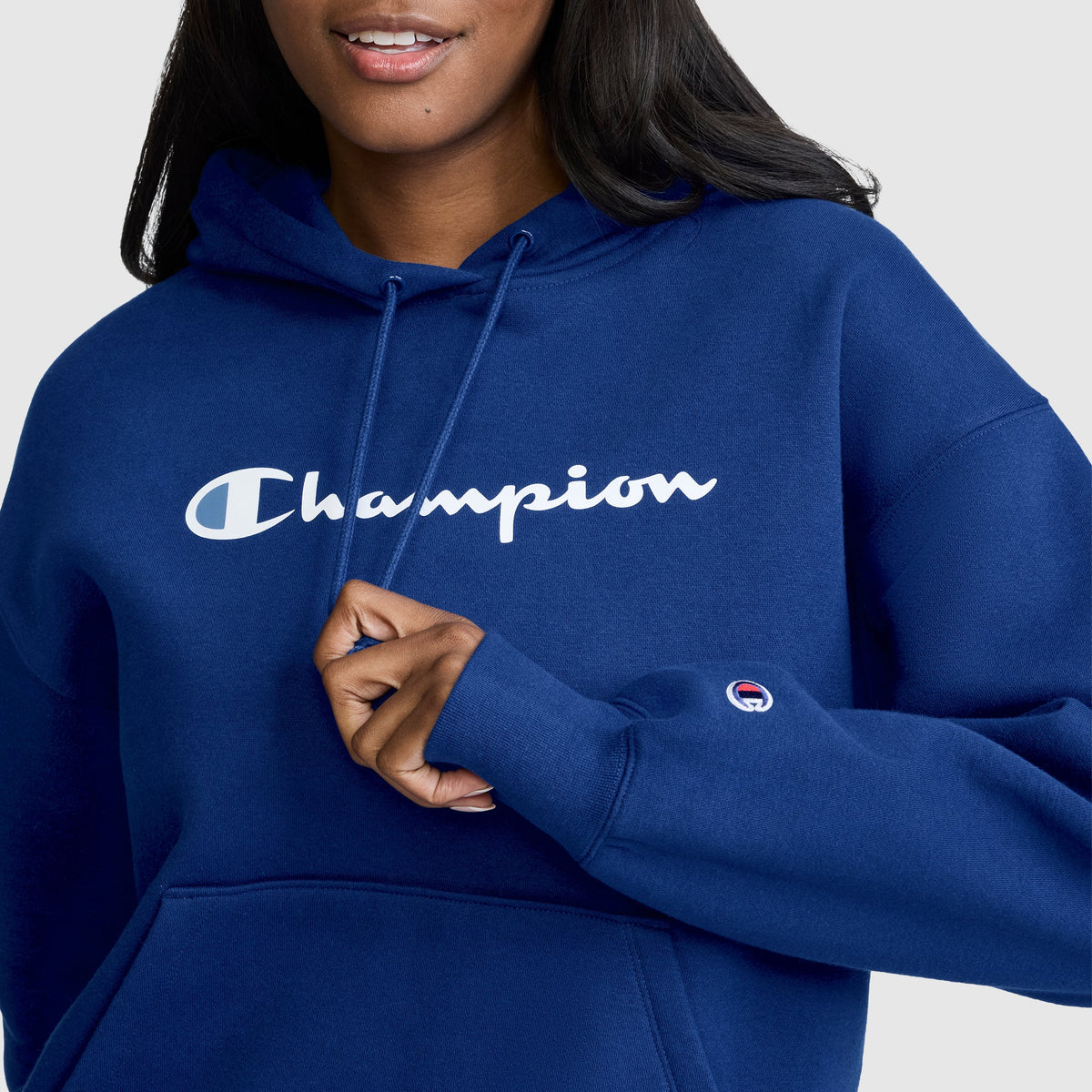 A person wearing a blue Champion Women’s Powerblend Relaxed Hoodie, Script Logo holds the hood’s drawstring. This women’s hoodie by Champion features the script logo printed in white across the chest and is crafted from midweight fleece for added comfort.