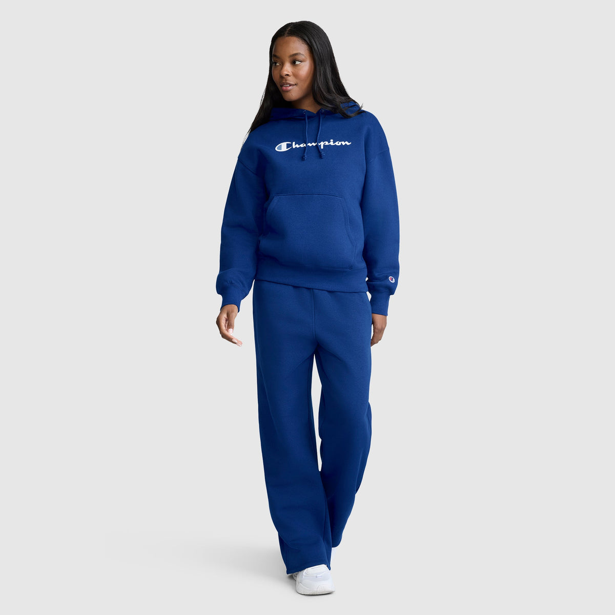 A person is wearing a blue Champion Women’s Powerblend Relaxed Hoodie with a script logo, made from cozy midweight fleece. They have paired it with matching blue sweatpants and white sneakers, standing and looking to the side against a plain light gray background.