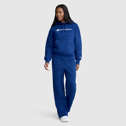 A person is wearing a blue Champion Women’s Powerblend Relaxed Hoodie with a script logo, made from cozy midweight fleece. They have paired it with matching blue sweatpants and white sneakers, standing and looking to the side against a plain light gray background.