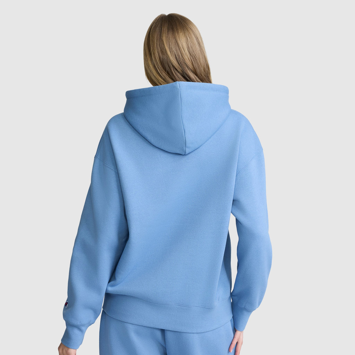 A person with long light brown hair is shown from the back, wearing a Champion Women’s Powerblend Relaxed Hoodie with a script logo and matching fleece sweatpants against a plain light background.