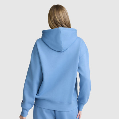 A person with long light brown hair is shown from the back, wearing a Champion Women’s Powerblend Relaxed Hoodie with a script logo and matching fleece sweatpants against a plain light background.