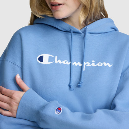 A person wearing a light blue Champion Women’s Powerblend Relaxed Hoodie, featuring the white script logo across the chest and a logo patch on the sleeve, stands with arms crossed in a relaxed pose, only the lower half of their face visible.