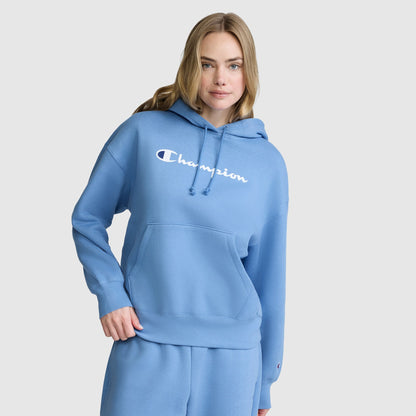 A woman wearing a light blue Champion Women’s Powerblend Relaxed Hoodie with a script logo and matching sweatpants, both crafted from cozy Powerblend fleece, stands against a plain white background.