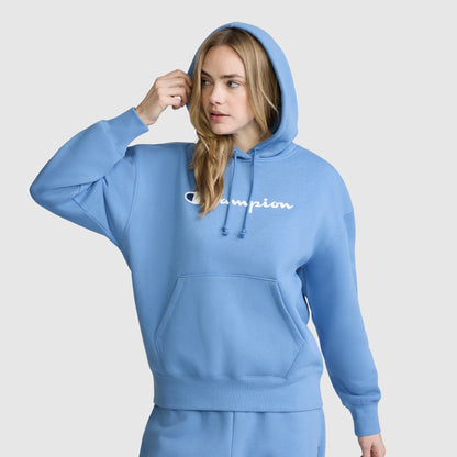 A woman wearing a light blue Champion Women’s Powerblend Relaxed Hoodie with a script logo stands against a plain background, touching her hair with one hand and looking to the side.