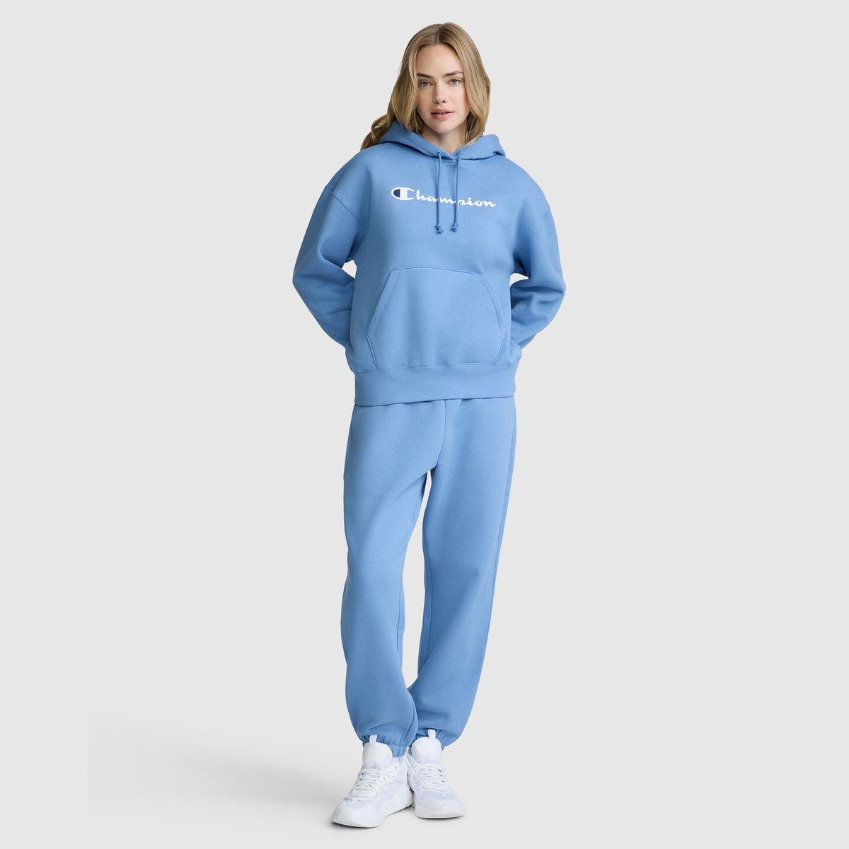 A person is seen facing forward against a plain background, wearing the Champion Women’s Powerblend Relaxed Hoodie, Script Logo by Champion in light blue, along with matching sweatpants and white sneakers. The hoodie, made from soft Powerblend fleece, features a relaxed fit and the individual has their hands in the hoodie pocket.