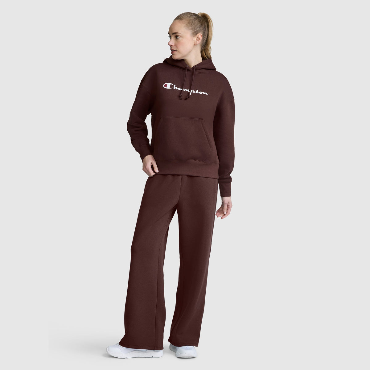 A person wearing a brown Champion Women’s Powerblend Relaxed Hoodie with a script logo and matching pants stands against a light gray background, looking to the side. Their light hair is styled in a ponytail, and they finish the outfit with white sneakers. The relaxed fit and soft Powerblend fleece from Champion lend comfort and effortless style to this casual look.