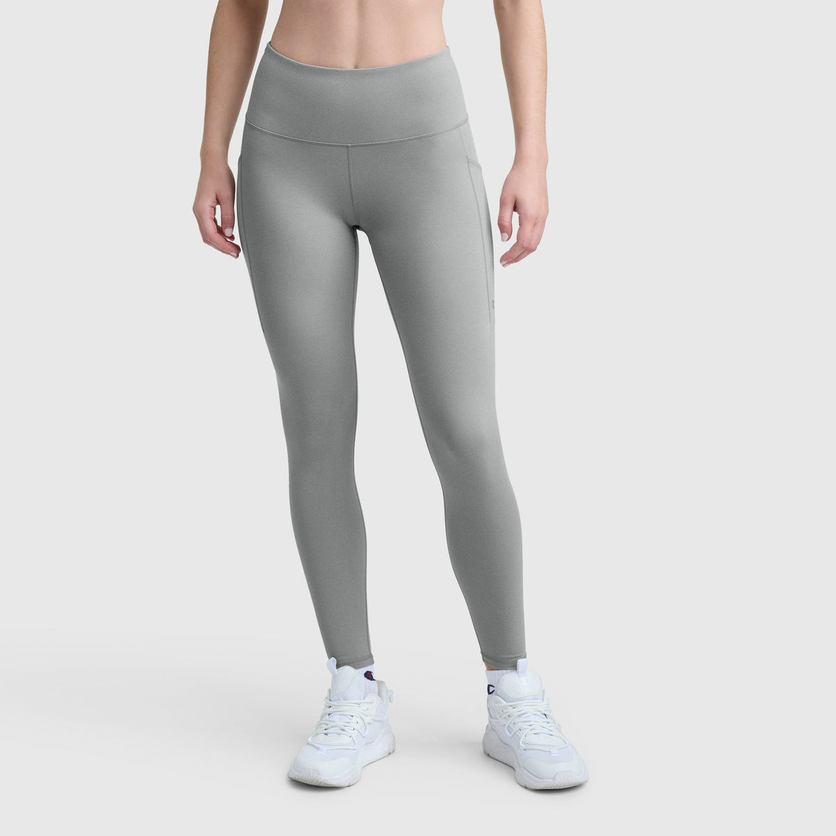 A person wears Champion C Logo light gray, high-waisted stretch leggings with pockets and white athletic shoes against a plain light background. The silky soft leggings have a smooth finish. The person's head is not visible.