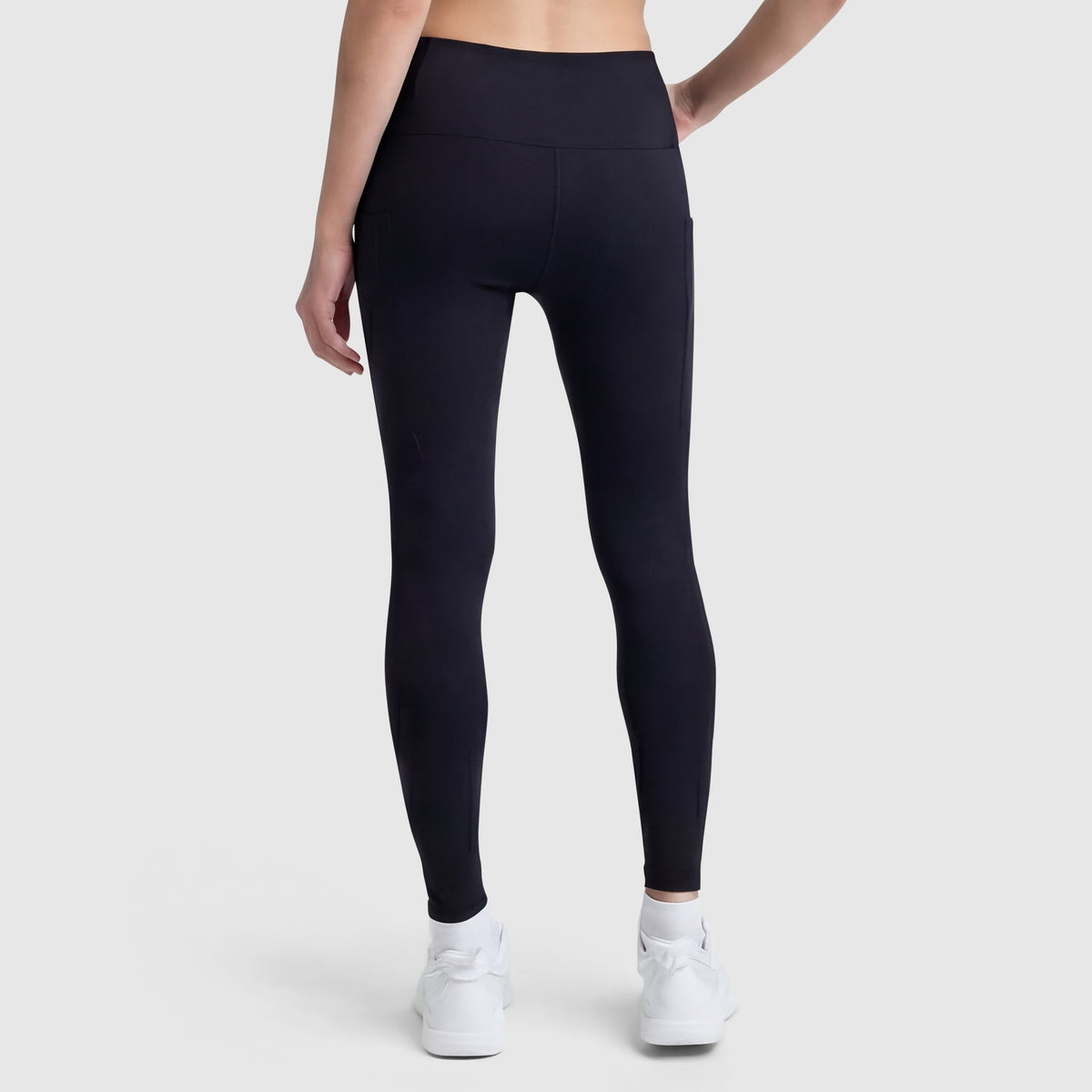 A person wearing Champion C Logo black high-waisted leggings with side pockets and white sneakers stands facing away, one hand on their hip, against a plain light gray background.