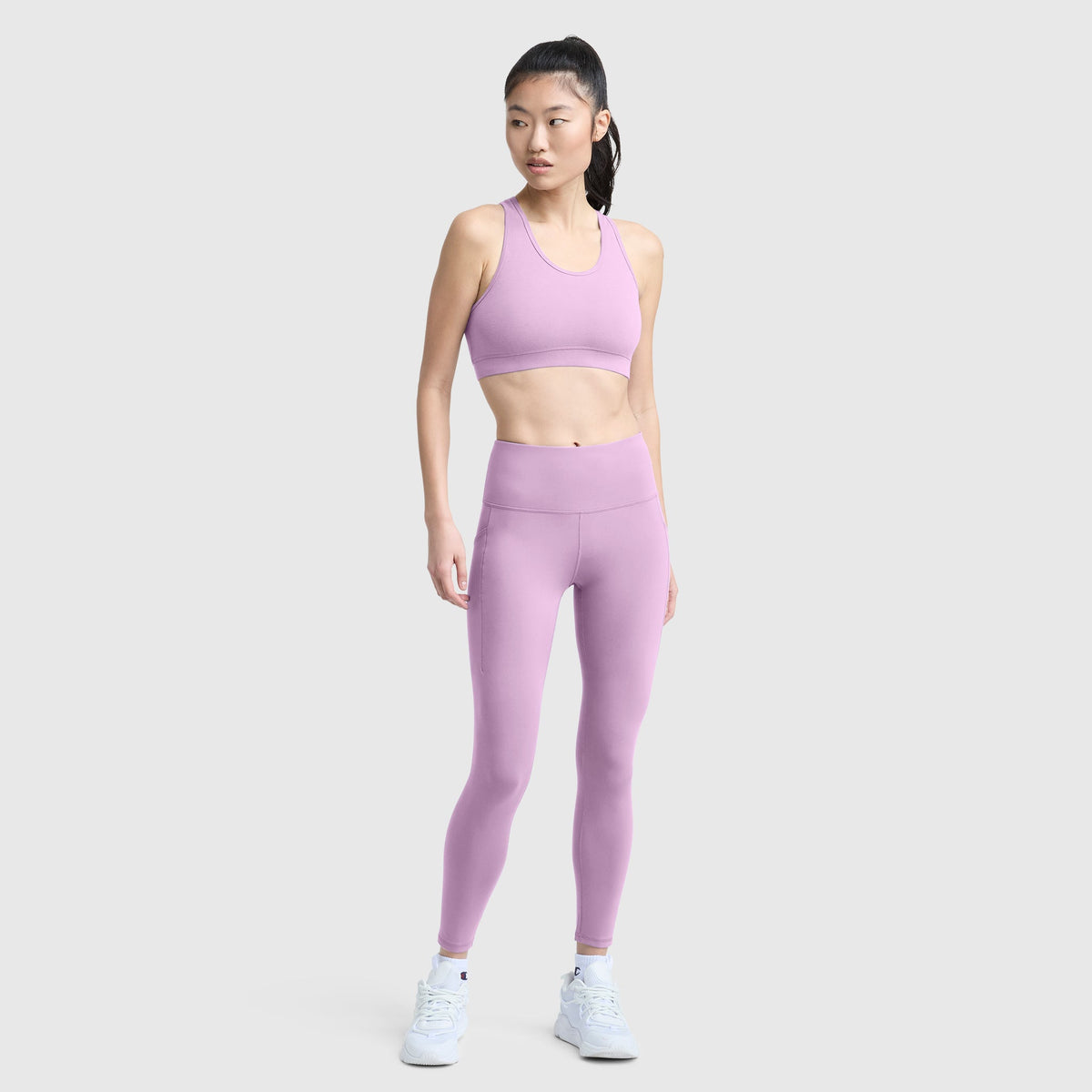 A woman stands against a plain light gray background, facing slightly to the side with her hands by her sides, wearing Champion’s C Logo leggings in light purple with pockets and a matching sports bra. She completes the look with white sneakers and a ponytail hairstyle.