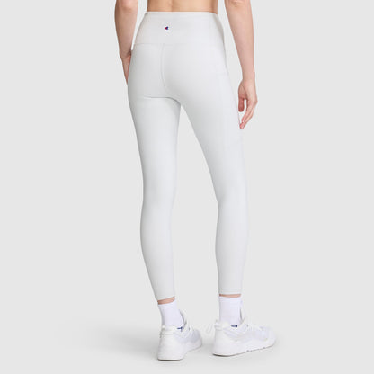 A person is wearing Champion’s Leggings, C Logo—high-waisted leggings crafted from soft peached fabric with side seams, visible stitching, discreet side pockets, and a small Champion logo on the waistband—paired with white sneakers and socks, standing against a plain light background.