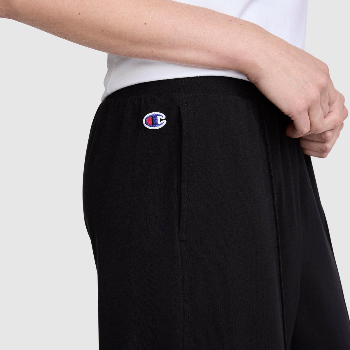 A person is seen wearing Champion Open Bottom Pants in black stretch jersey with the signature logo near the pocket, styled with a white shirt. The image focuses on the midsection, arm, and hand against a plain background, showcasing the athletic design of these Champion pants.