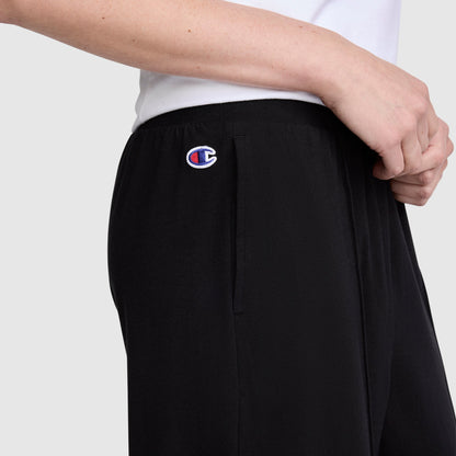 A person is seen wearing Champion Open Bottom Pants in black stretch jersey with the signature logo near the pocket, styled with a white shirt. The image focuses on the midsection, arm, and hand against a plain background, showcasing the athletic design of these Champion pants.