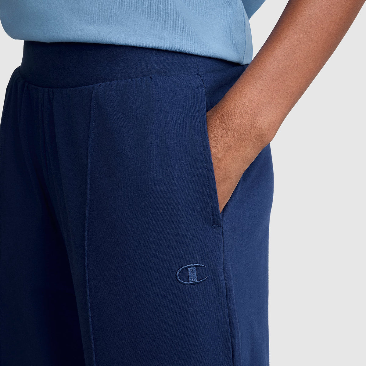 A person wearing the Champion Open Bottom Pant in navy blue, styled like stretch jersey pants with an embroidered logo on the thigh and a hand in the pocket. The person is also wearing a light blue shirt.