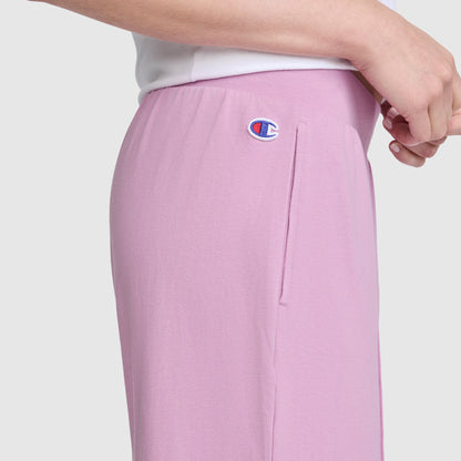 A person is shown from the waist down in a side view, wearing light pink Champion Open Bottom Pant and a white top, with one hand resting near their hip.