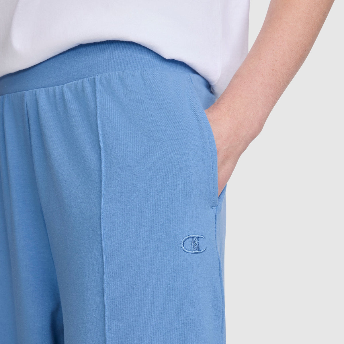 A person is wearing light blue Champion Open Bottom Pant stretch jersey athletic pants, featuring the Champion logo and with one hand in the pocket. The pants are paired with a white shirt, and only the lower torso and part of one arm are visible.