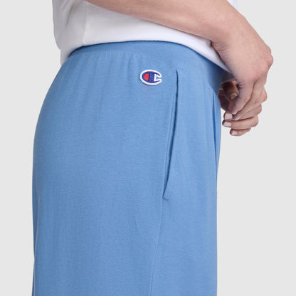 A person wearing Champion Open Bottom Pants in light blue stretch jersey fabric and a white shirt, with one hand near the side pocket. Only the lower torso and part of the arm are visible against a plain background, capturing the relaxed comfort of Champion’s athletic wear.