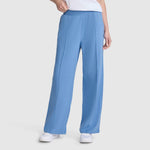 A person wearing Champion’s Open Bottom Pant—loose-fitting, light blue athletic pants made from comfortable stretch jersey with an elastic waistband—paired with white sneakers and a white top, stands against a plain light gray background.