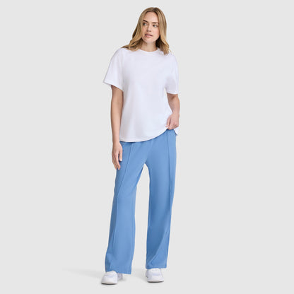 A woman with light hair poses casually against a plain background, wearing a white short-sleeve t-shirt, the Champion Open Bottom Pant in light blue made from stretch jersey, and white sneakers. She has one hand in her pocket for a relaxed look.