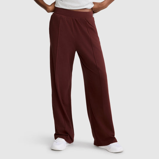 A person wears Champion’s Open Bottom Pant—a high-waisted, wide-leg maroon stretch jersey style with an athletic silhouette—paired with a tucked white shirt and white sneakers, standing against a plain light gray background. Only the lower body is visible.