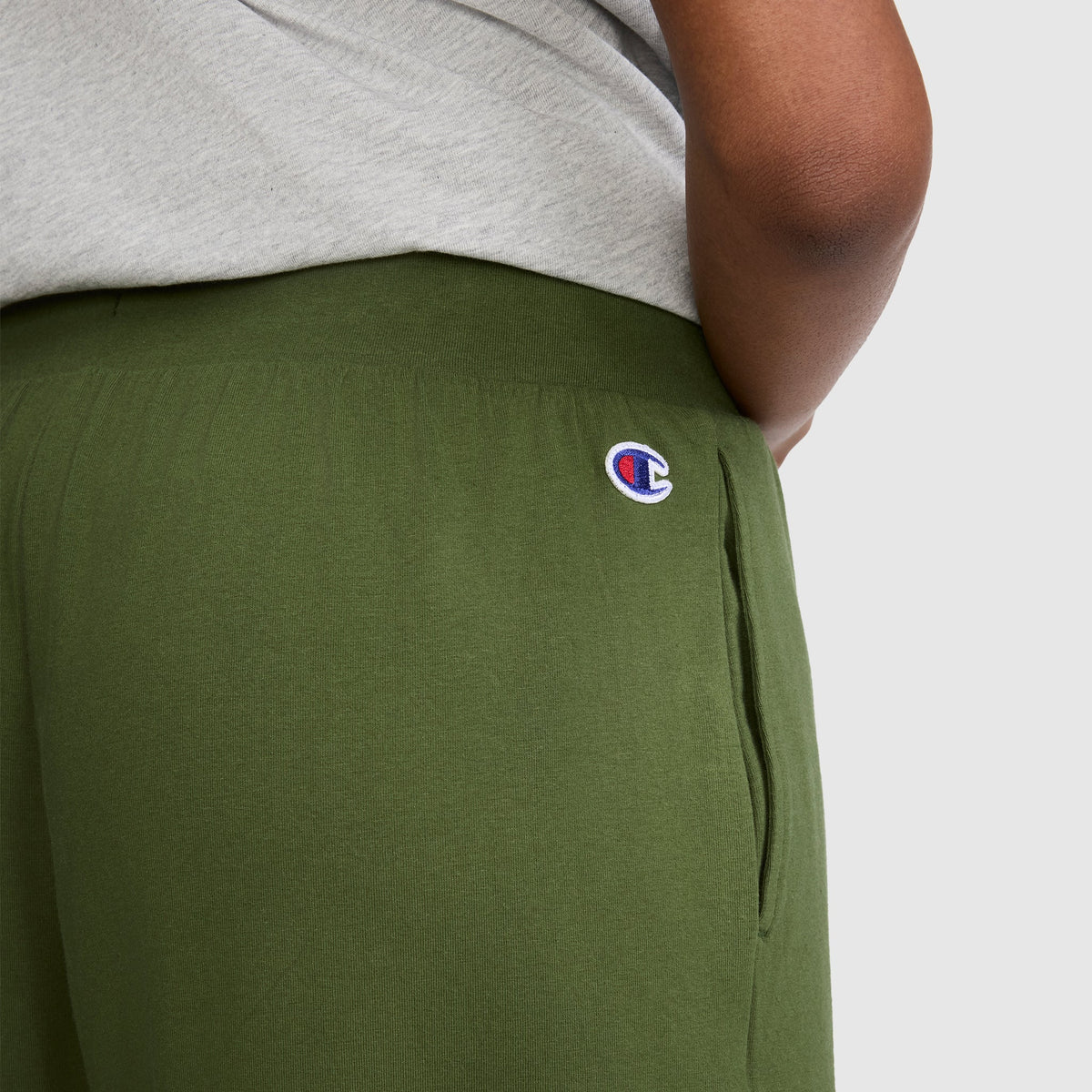 A person wearing Champion Open Bottom Pants in olive green, featuring the signature logo on the hip, paired with a light gray shirt. Only part of the torso and arm are visible, emphasizing the comfort and style of these Champion pants.