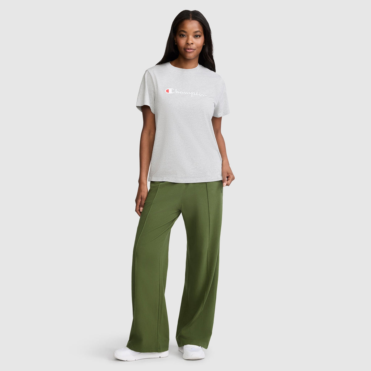 A woman stands smiling against a plain light gray background, wearing a light gray Champion t-shirt, white sneakers, and the Champion Open Bottom Pant—a loose-fit style in olive green stretch jersey.