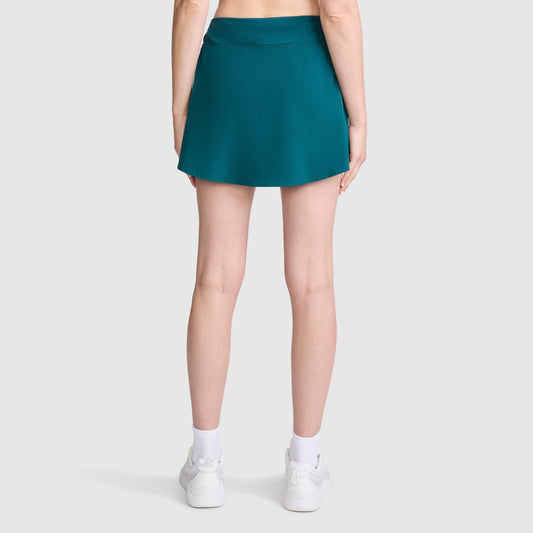 A person stands facing away from the camera against a plain light gray background, wearing the Champion Courtside Skort, C Logo in teal, paired with white socks and white sneakers.