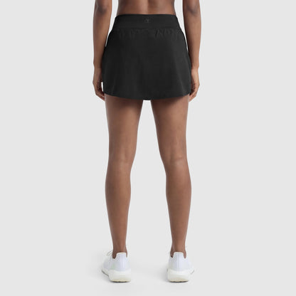 Someone stands with their back to the camera, displaying Champion’s Courtside Skort, C Logo in black along with white athletic shoes against a simple light background. Only the sporty court-inspired lower body is shown.