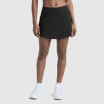 A person models Champion’s Courtside Skort, C Logo in black, styled with white socks and sneakers against a plain light gray background. Only the lower torso and legs are shown.