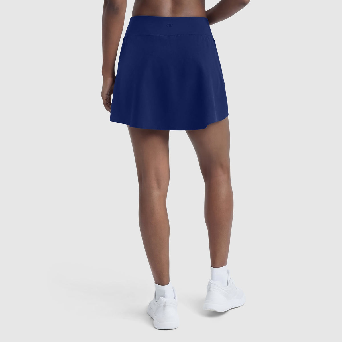 A person in a Champion Courtside Skort with C Logo, white socks, and white sneakers stands facing away from the camera against a plain light background, highlighting modern court style.