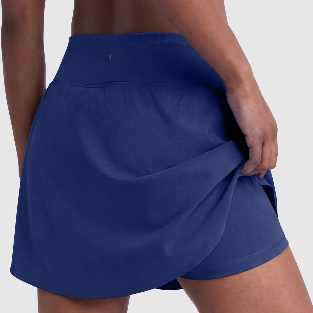 A person in a Champion Courtside Skort (C Logo) lifts the outer layer to reveal matching blue shorts, highlighting classic court style against a plain, light background.