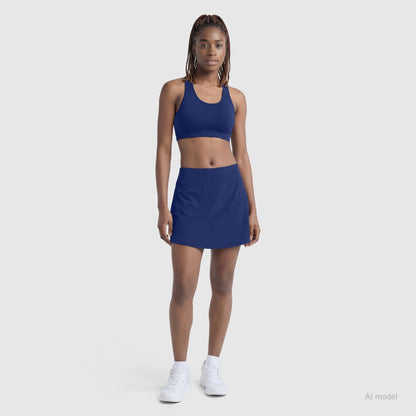 A woman with braided hair stands facing forward in the Champion Courtside Skort (C Logo) and matching sports bra, paired with white sneakers. She poses against a light gray background, showcasing a classic court style look.
