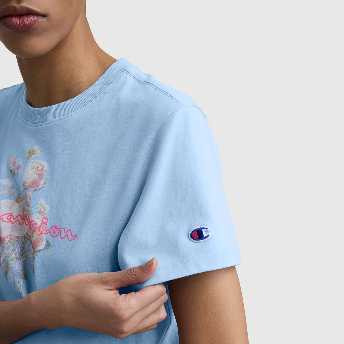 A person wears a light blue Champion Blurred Roses Graphic T-Shirt with a floral graphic and cursive text on the front, made from combed compact cotton, and featuring a small C logo patch on the sleeve, shown against a plain background.