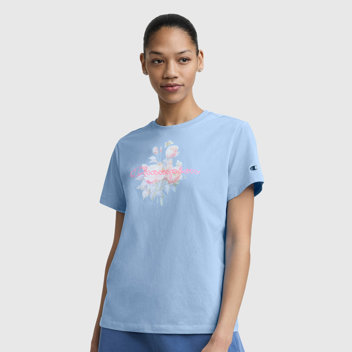 A woman in a light blue Champion Blurred Roses Graphic T-Shirt with C Logo, made from combed compact cotton, stands against a plain white background. Her hair is tied back and she gazes forward with a soft expression.