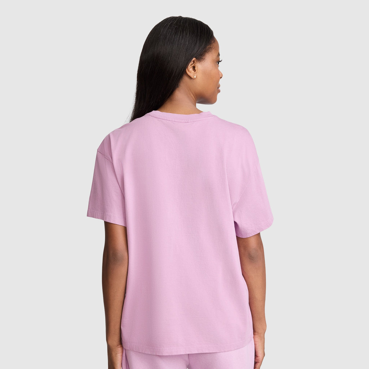 A person with long dark hair is standing facing away, wearing the Champion Short Sleeve Loose Fit T-Shirt in light pink and matching pants against a plain light gray background.
