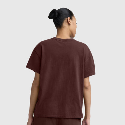 A person with dark hair in a bun is seen from the back, wearing the Champion Short Sleeve Loose Fit T-Shirt in brown along with matching pants, standing against a plain light gray background.