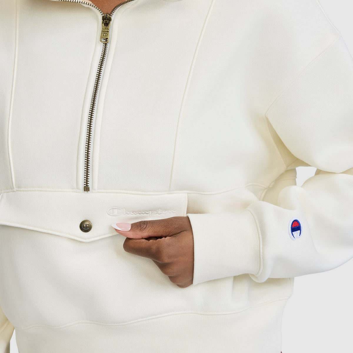 A person wearing an off-white Champion Training Half-Zip Fleece Hoodie, with a close-up of their hand near the snap button pocket. The Champion logo patch is visible on the sleeve, emphasizing this stylish athletic pullover.