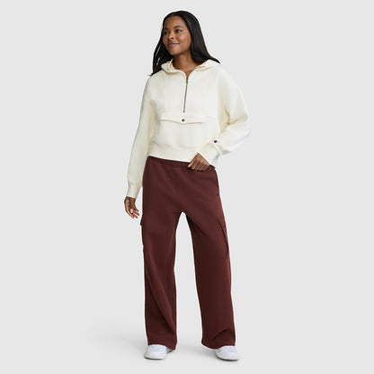 A woman wearing a white Champion Training Half-Zip Fleece Hoodie and loose maroon cargo pants stands smiling with one arm slightly extended against a plain light gray background. She is also wearing white shoes.