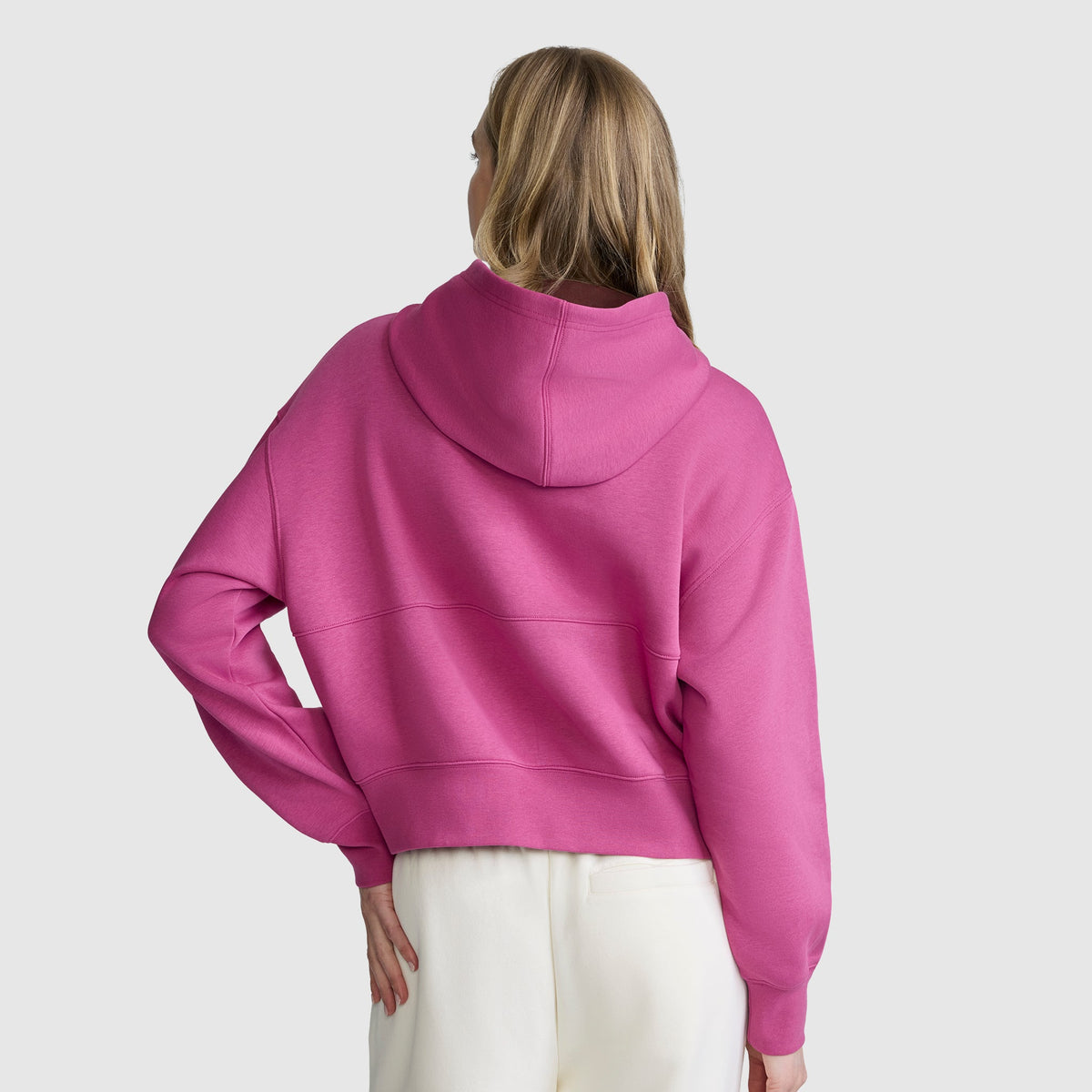 A person with long blonde hair is wearing a bright pink Champion Training Half-Zip Fleece Hoodie and white pants, standing with their back to the camera against a plain light gray background.