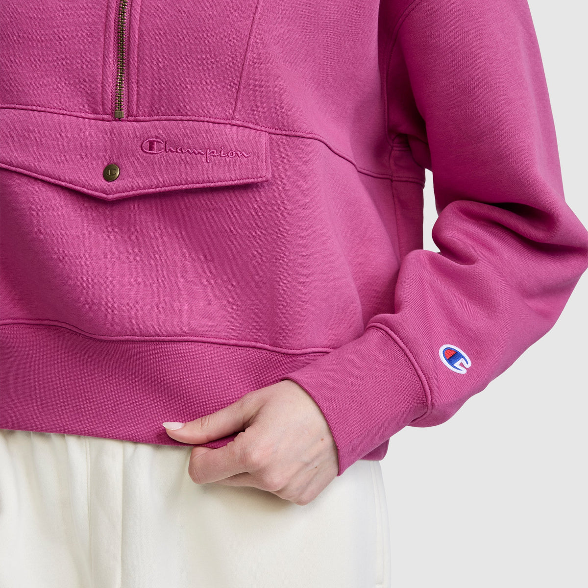 A person wearing a pink Champion Training Half-Zip Fleece Hoodie with a front pocket and white pants, hand resting near the pocket. The Champion logo is visible on the sleeve and above the pocket.
