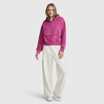 A woman stands against a plain light gray background wearing a bright pink Champion Training Half-Zip Fleece Hoodie with a front pocket, paired with white wide-leg pants and white sneakers.