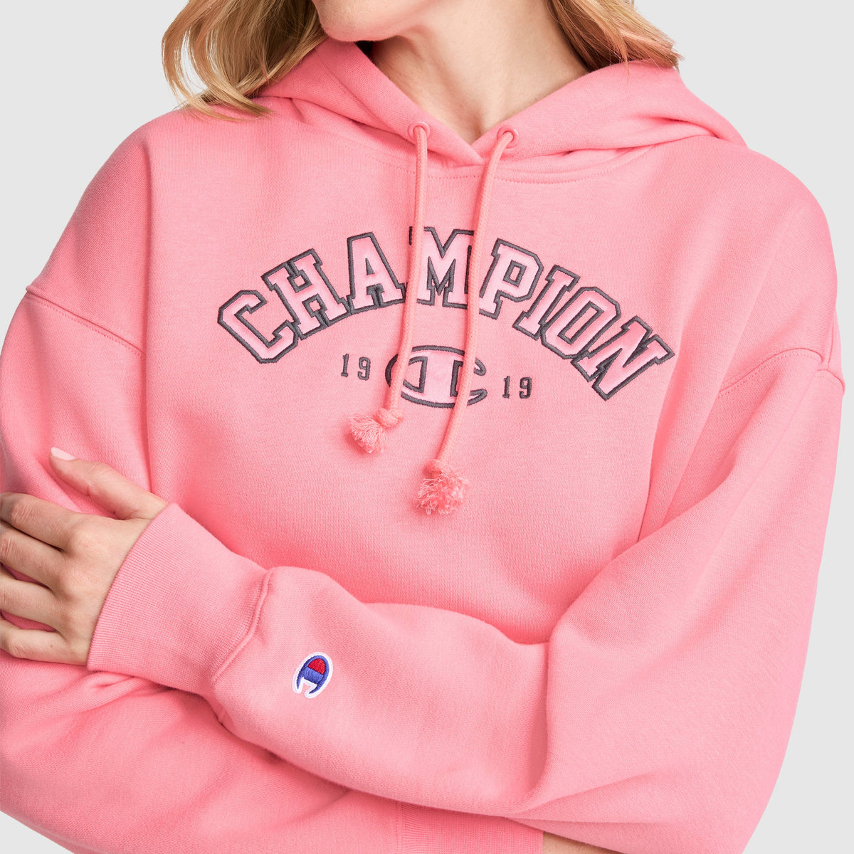 A person wearing a pink Champion Fleece Hoodie, Satin Applique with "CHAMPION 1919" on the front and arms crossed, displaying the Champion logo on the sleeve. This varsity-style hoodie from Champion features classic details and stands out with its bold design.