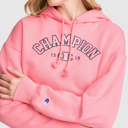 A person wearing a pink Champion Fleece Hoodie, Satin Applique with "CHAMPION 1919" on the front and arms crossed, displaying the Champion logo on the sleeve. This varsity-style hoodie from Champion features classic details and stands out with its bold design.