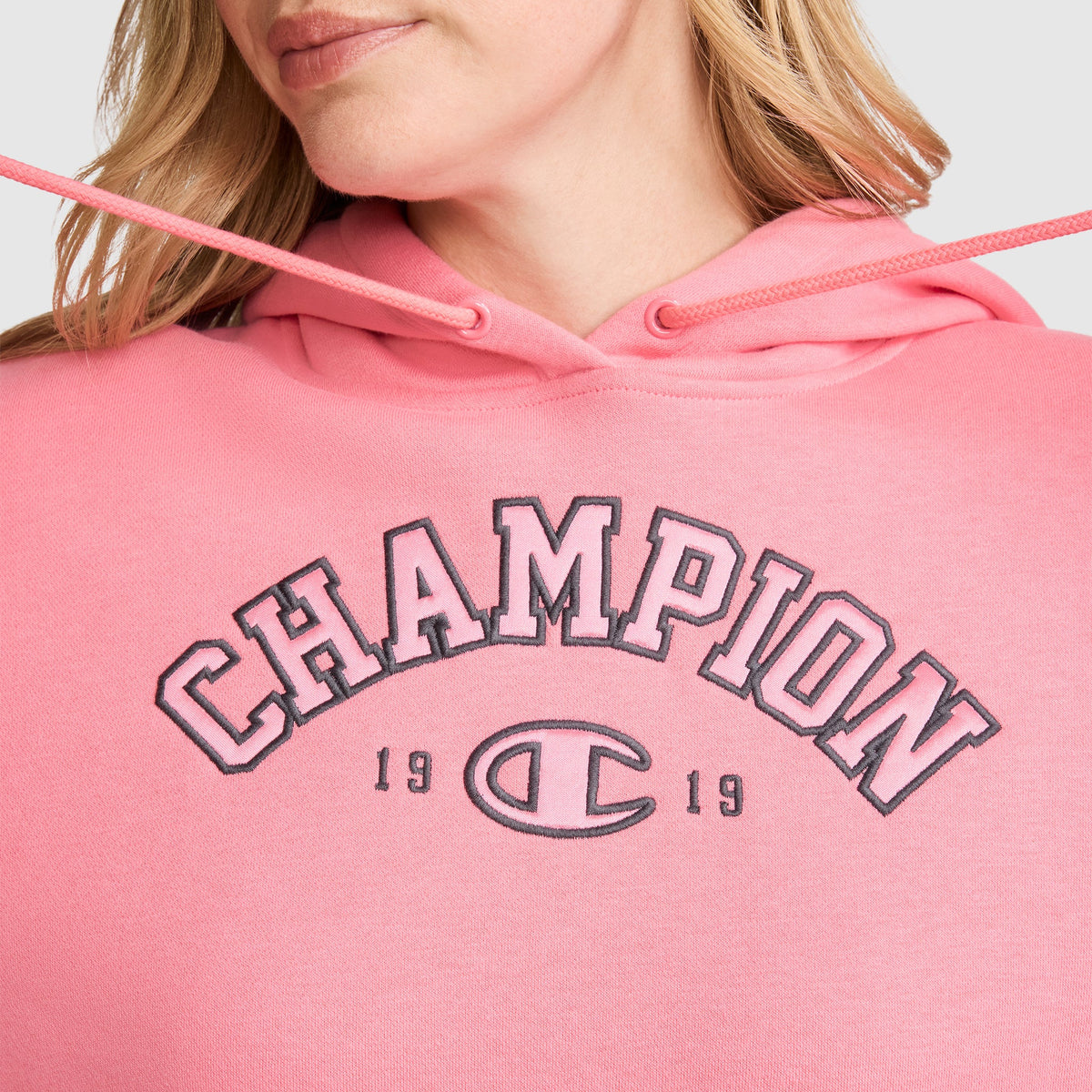 A person is wearing a Champion Fleece Hoodie, Satin Applique in pink with "CHAMPION 1919" and the brand logo on the chest. Only their lower face, neck, and upper torso are visible.