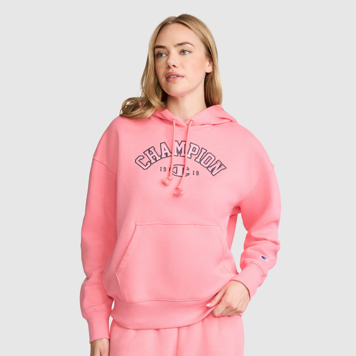 A woman stands against a plain background with one hand in her pocket, wearing the Champion Fleece Hoodie, Satin Applique and matching pants, effortlessly showcasing a modern take on varsity style while looking slightly to the side.