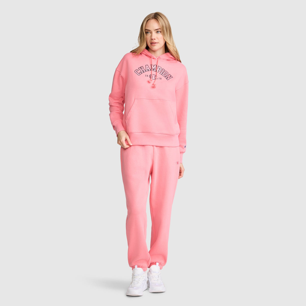 A woman stands facing forward against a plain light gray background, wearing a matching pink Champion Fleece Hoodie, Satin Applique, and sweatpants with white sneakers. The varsity-style hoodie by Champion features a subtle satin applique, adding a trendy touch to her sporty look.