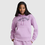 A woman stands against a plain light gray background, smiling with one hand in the pocket of her light purple Champion Fleece Hoodie, Satin Applique varsity style hoodie and matching pants.