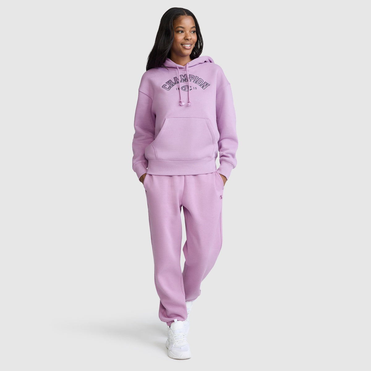 A person smiles while wearing a matching lavender Champion Fleece Hoodie, Satin Applique, and sweatpants set with white sneakers, standing against a plain white background.